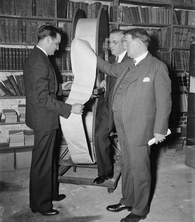Three men at the National Archives examine two stacked spools of Good Roads petitions taller than they are.