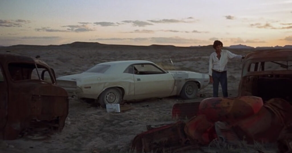 Film still of Kowalski contemplating inevitability while touching a rusted out vehicle hull on the side of Highway 50.