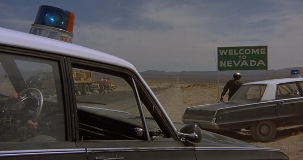 Film still of two police cars stopped, now revealing the road sign reads "Welcome to Nevada"