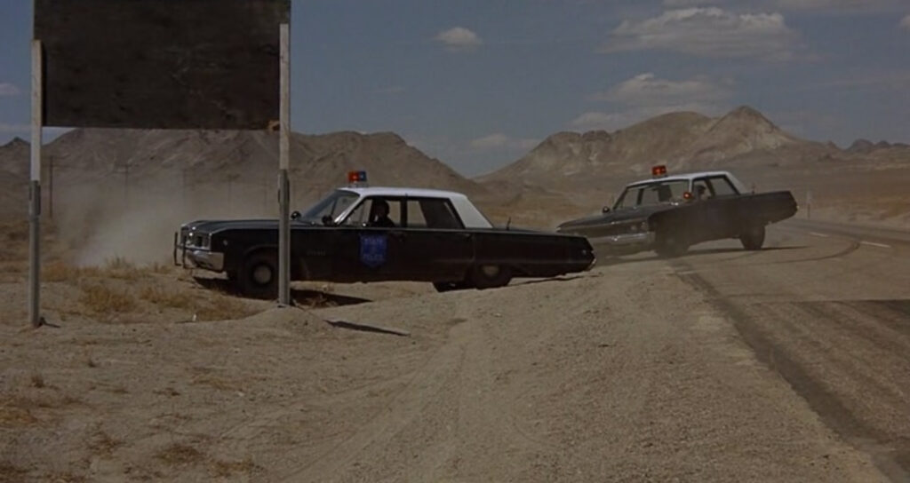 Two police cars halting their pursuit in front of a large sign facing away from the camera